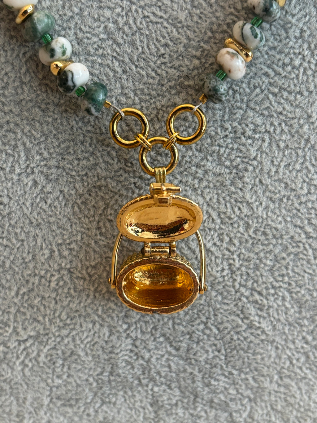 Gold locket charm on a beaded necklace against a textured gray background