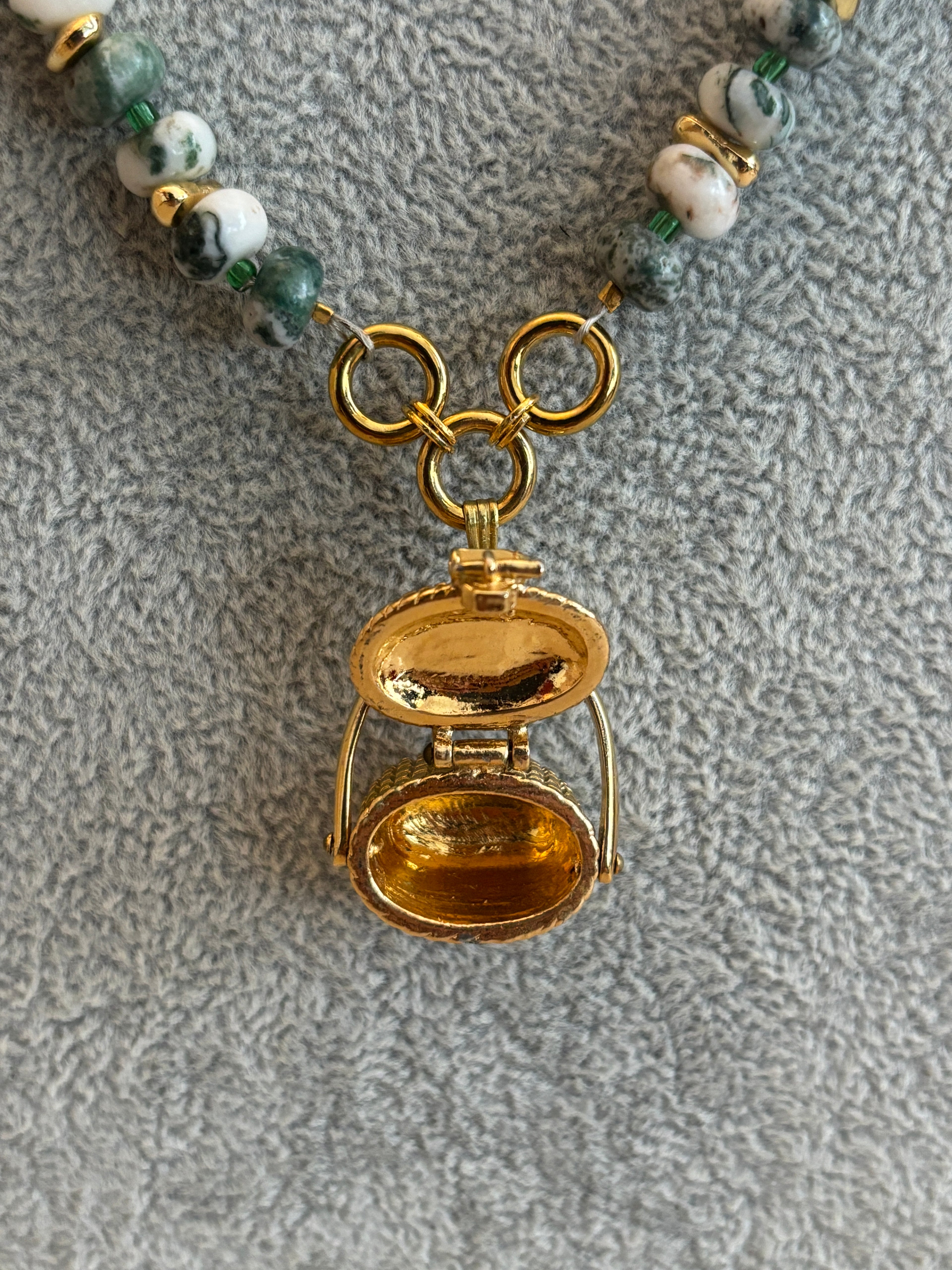 Gold locket charm on a beaded necklace against a textured gray background