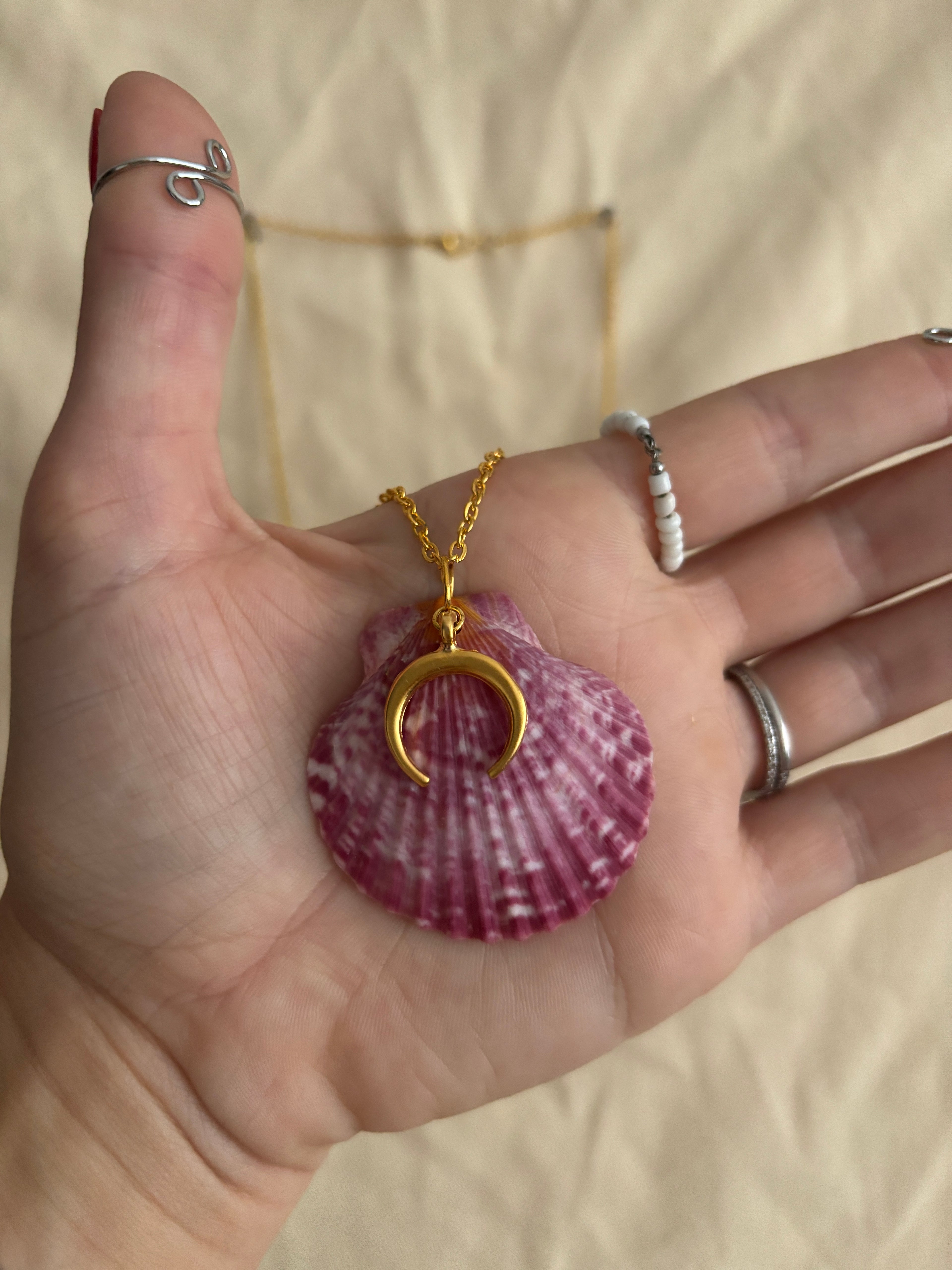Hand wearing a gold necklace with a shell pendant on a beige surface