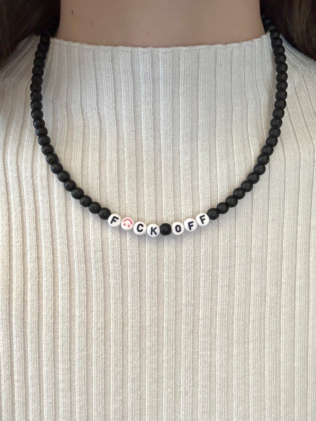 Necklace with beads spelling 'F*CK OFF' on a white ribbed sweater.