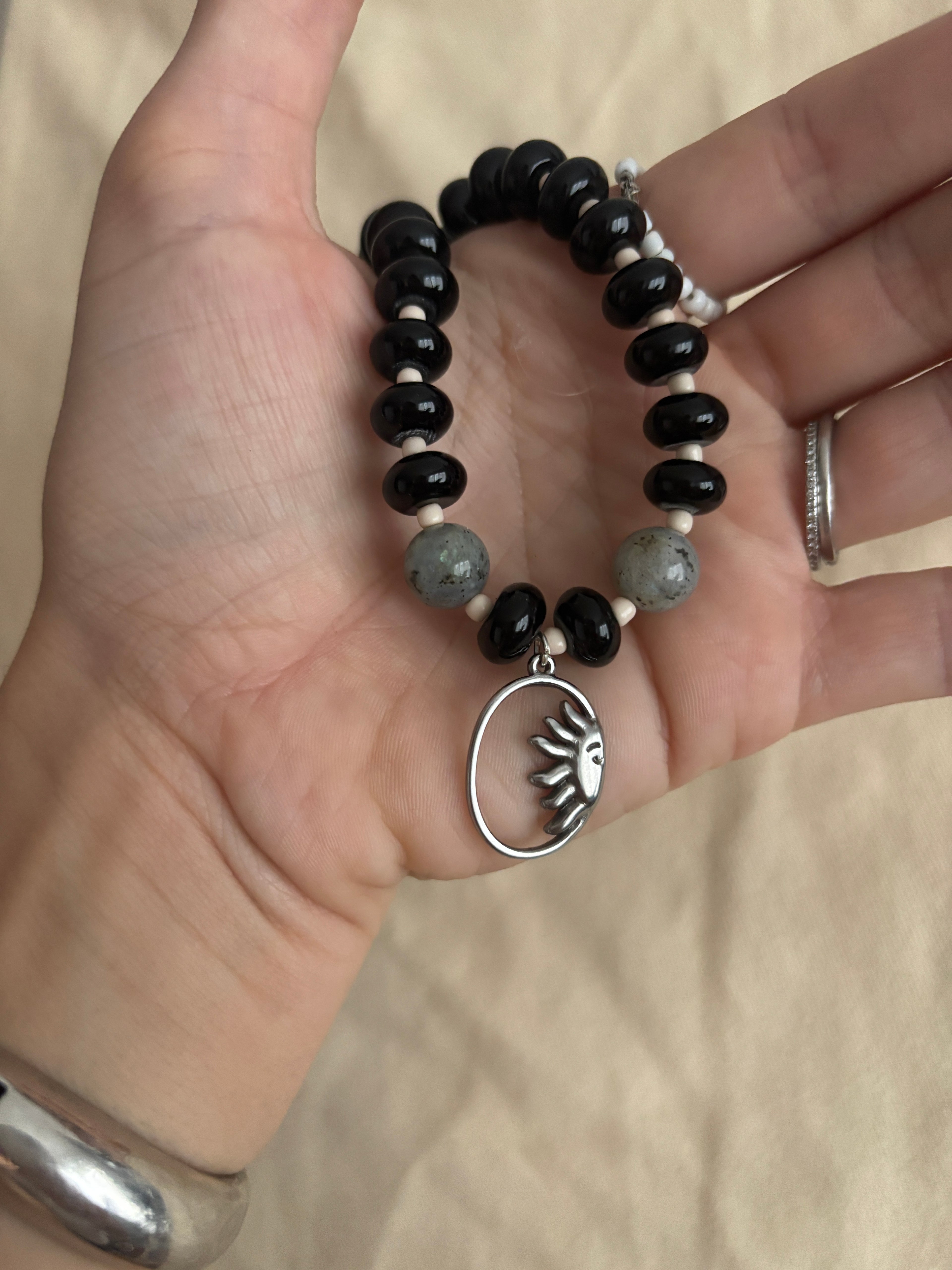 Black beaded bracelet with a silver sun charm held in a hand against a beige background