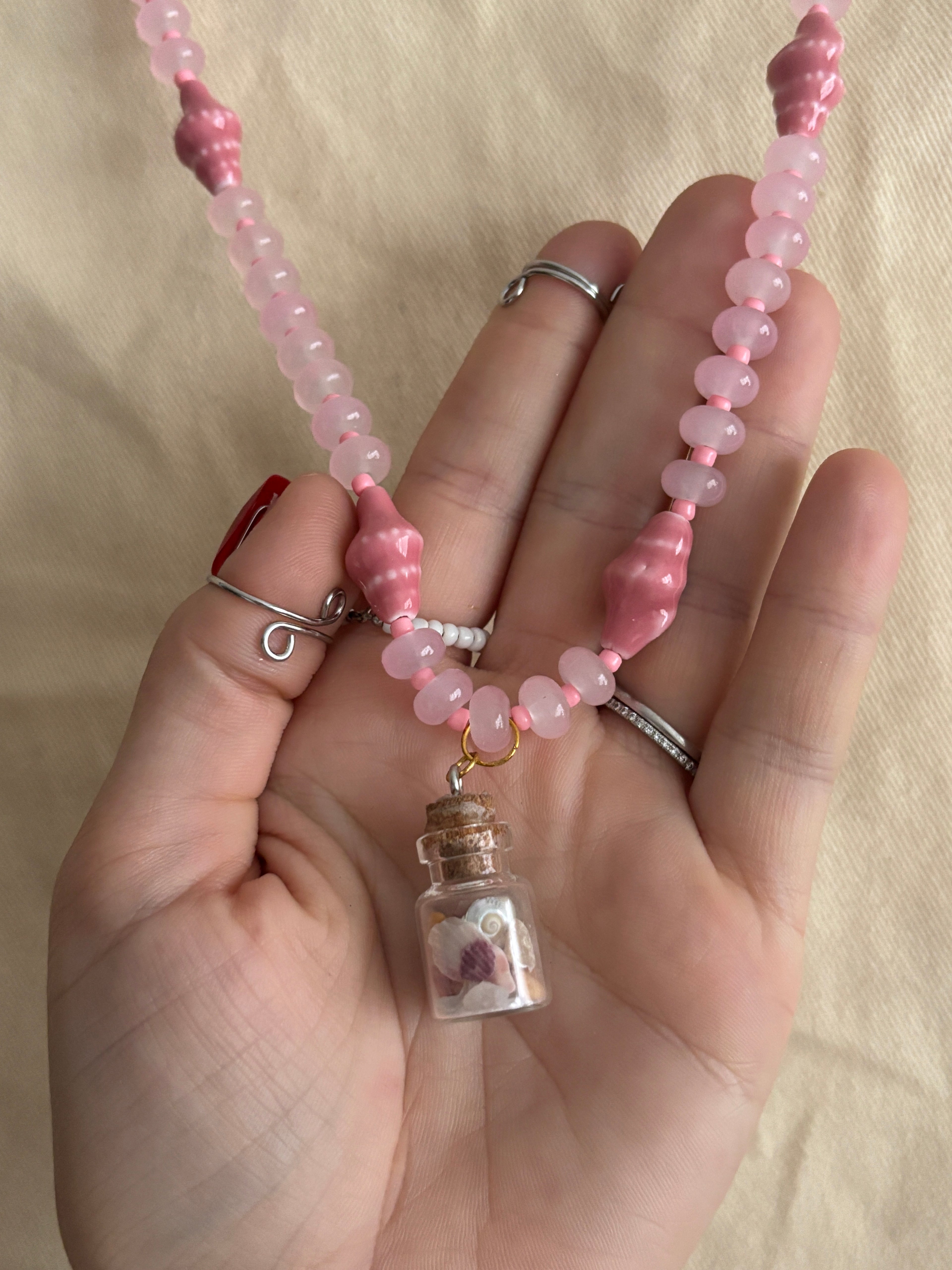 Hand holding a pink beaded necklace with a small jar charm against a beige background