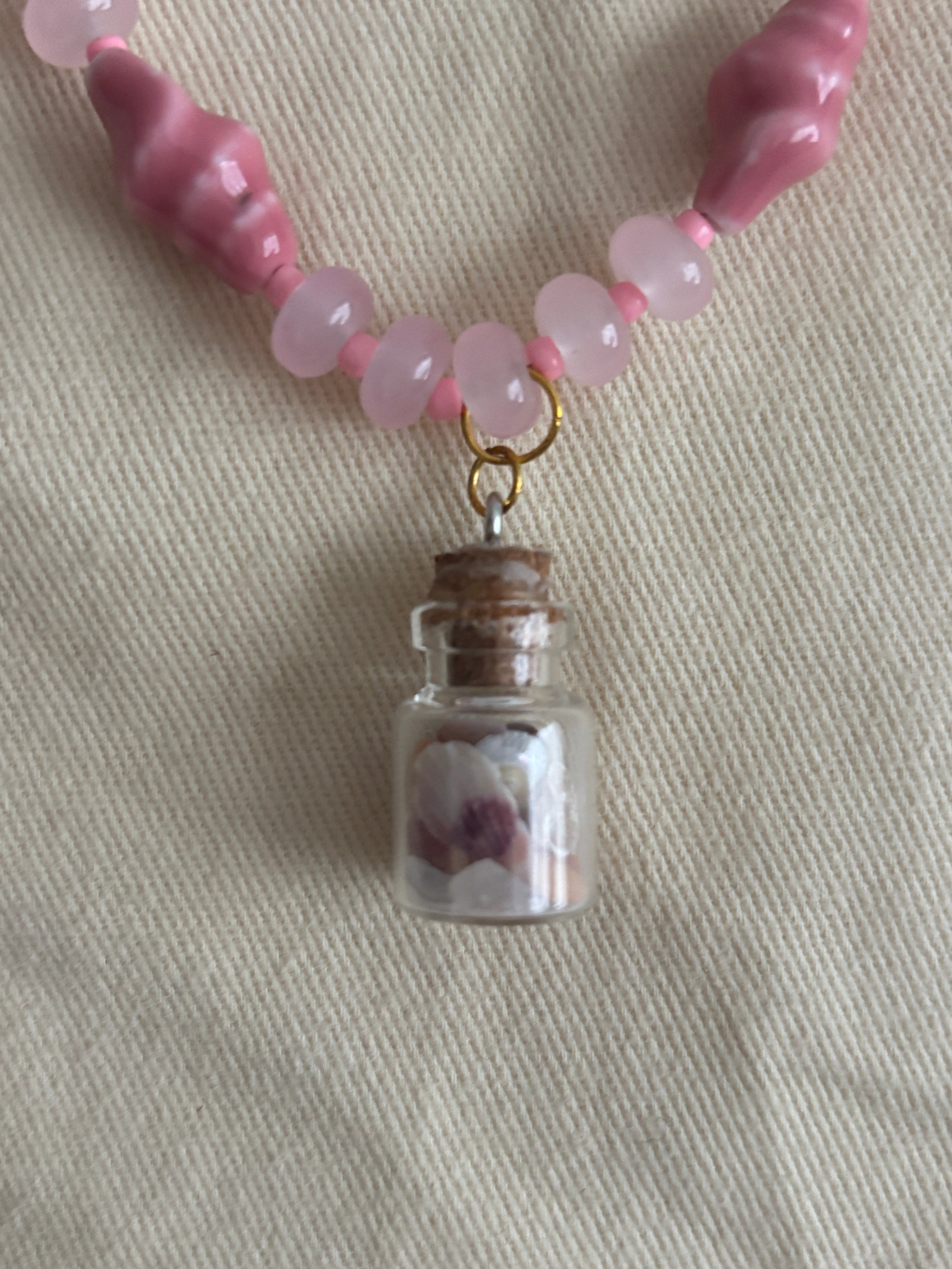 Pink beaded necklace with a small glass jar charm on a beige fabric background