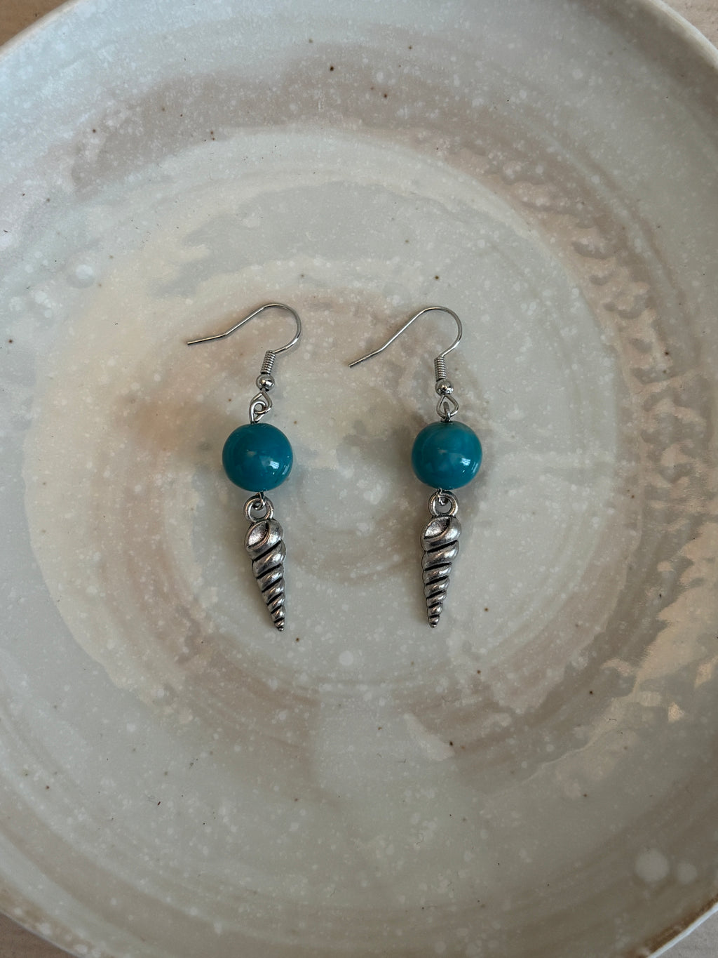 Pair of earrings with turquoise beads and silver shells on a textured ceramic plate.