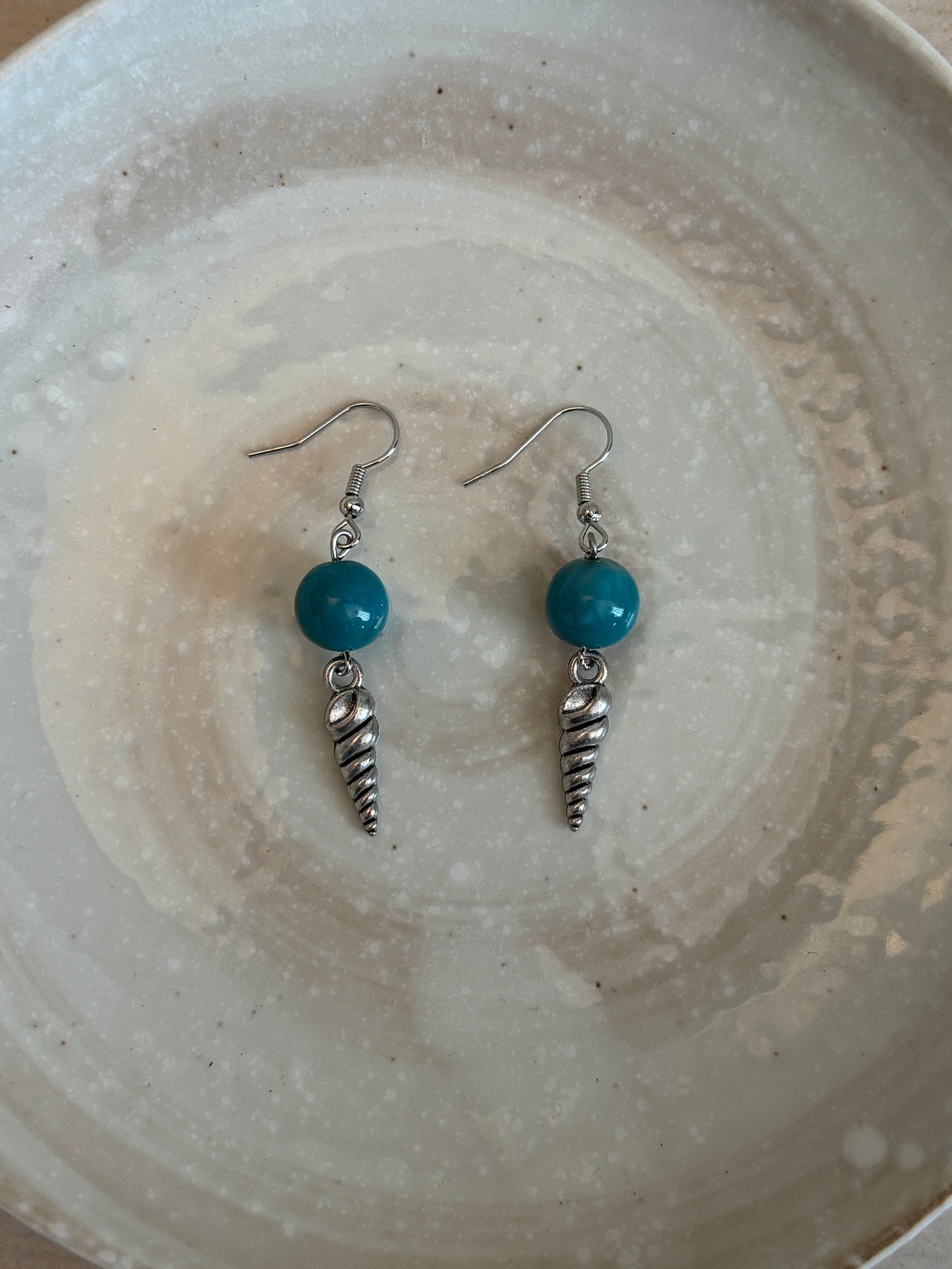 Pair of earrings with turquoise beads and silver shells on a textured ceramic plate.