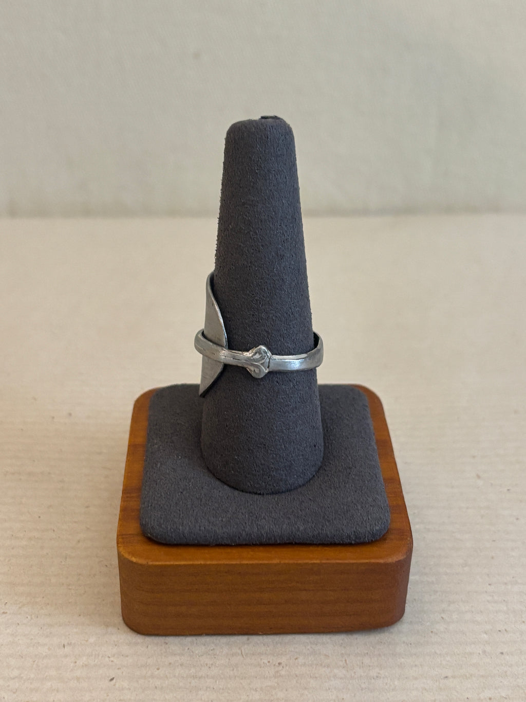 Ring holder with a wooden base and gray cushion holding a ring on a beige wall.